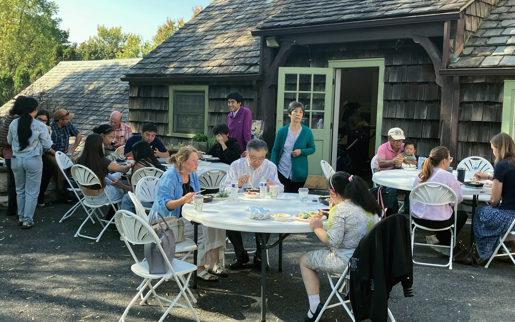 People eating together at folding tables outside a shingled hall after a service.